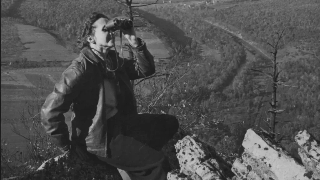 Rachel Carson em trabalho de campo, sentada num afloramento rochoso elevado, a observar a paisagem com binóculos, com um vale agrícola ao fundo.