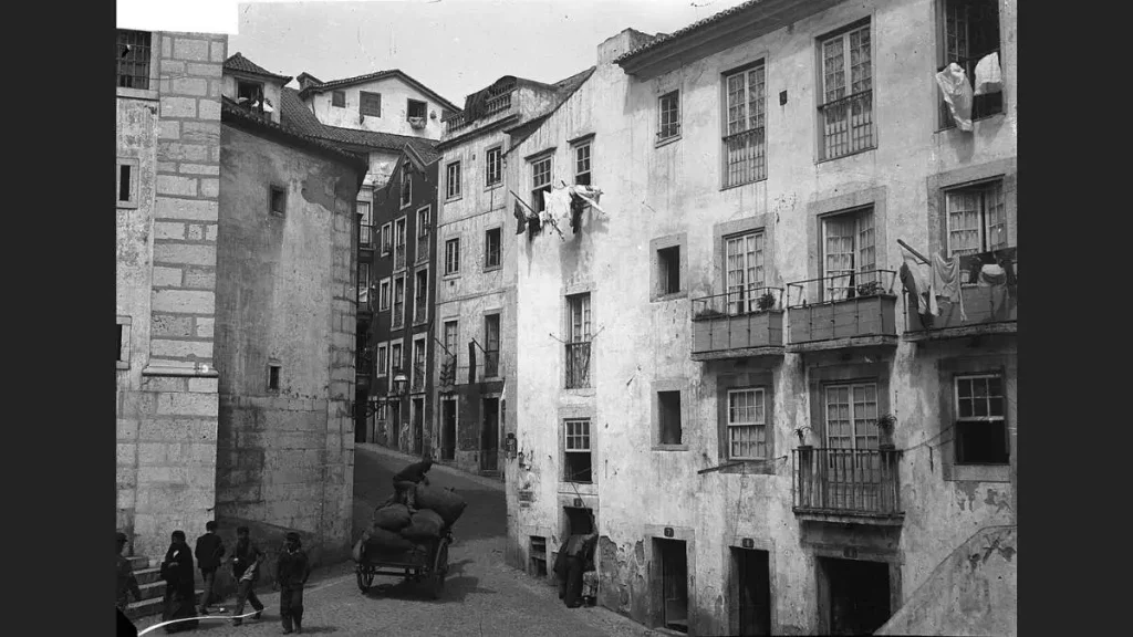 Largo de São Miguel, em Alfama, em fotografia de cerca de 1900: edifícios populares de várias alturas com janelas de guilhotina, roupa estendida em cordas e varandas, uma carroça com sacos ao centro do largo, figuras a passar

