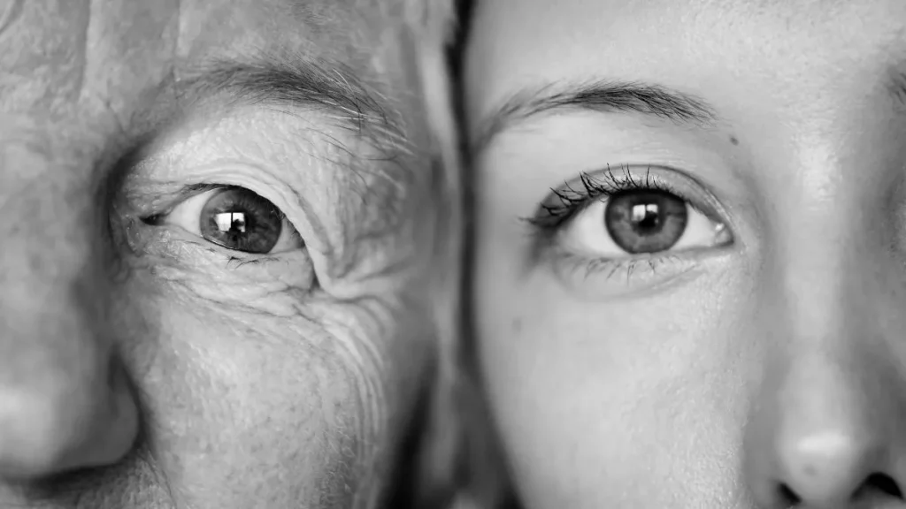 Plano aproximado a preto e branco de dois rostos lado a lado, uma mulher idosa e uma mulher jovem, com os olhos abertos a olhar para a câmara, contraste entre a pele enrugada de um e lisa do outro. Esperança de vida