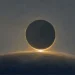 Ilustração a óleo de um eclipse do Sol, com o disco lunar escuro sobre um ponto de luz intensa no bordo inferior e um halo ténue em redor, acima de uma linha de horizonte curva