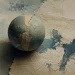 Esfera texturada em azul e creme pousada sobre uma folha de papel rasgada, com arcos de grelha cartográfica e manchas azuis que sugerem massas de terra