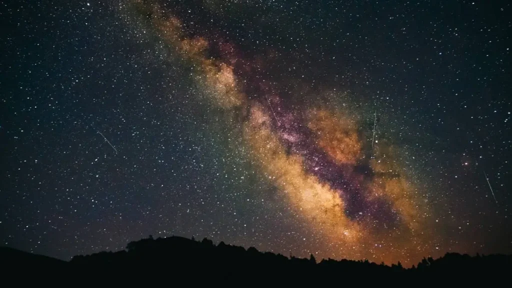 A faixa da Via Láctea na diagonal sobre uma serra em silhueta, com o núcleo galáctico em tons dourados e rastos de meteoros das perseidas
