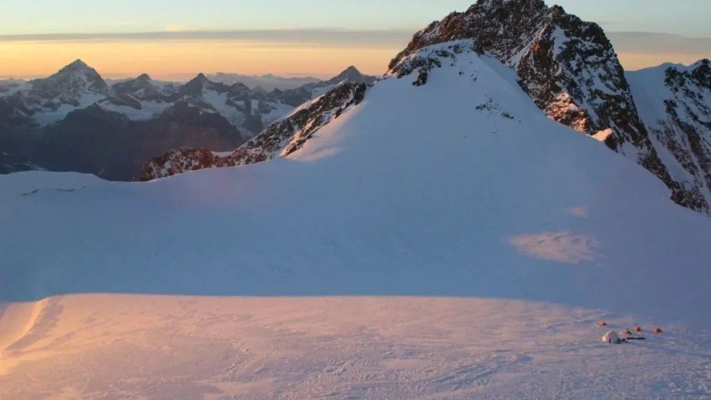 Planalto glaciar coberto de neve ao nascer do sol, com um pequeno acampamento de tendas laranja e uma cúpula branca no canto inferior direito, um pico rochoso à direita e uma cadeia de montanhas alpinas no horizonte alaranjado